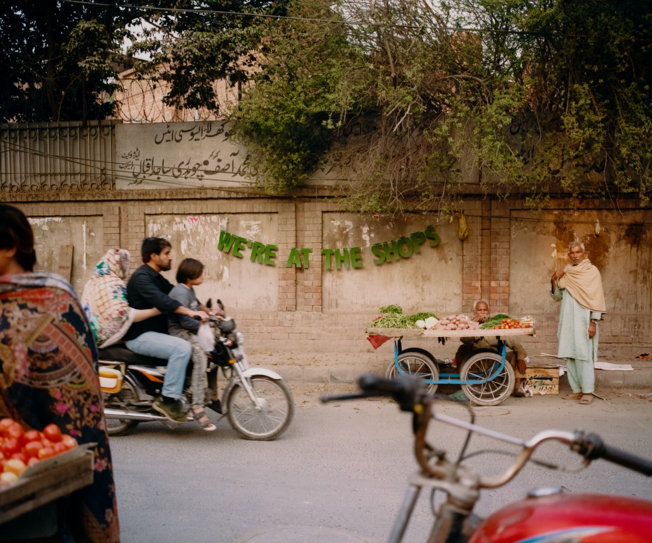 Three people ride on a motorcycle passing by fruit stands in the street.