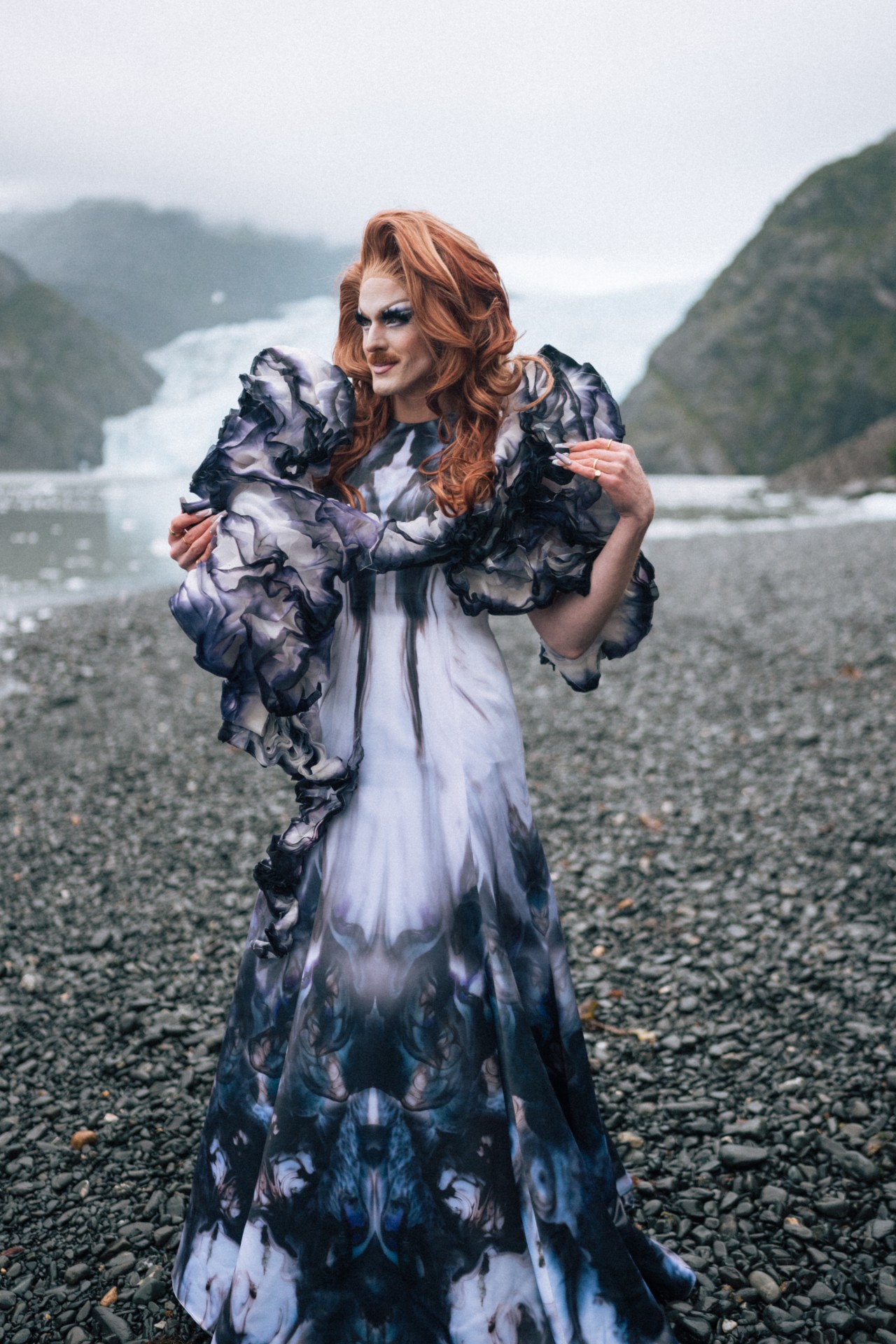 Pattie Gonia stands poses in front of a mountainous, icy landscape.