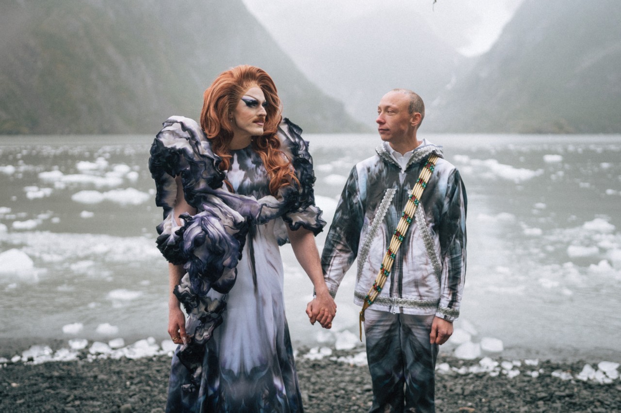 Pattie Gonia and Quinn Christopherson hold hands in front of an icy body of water.