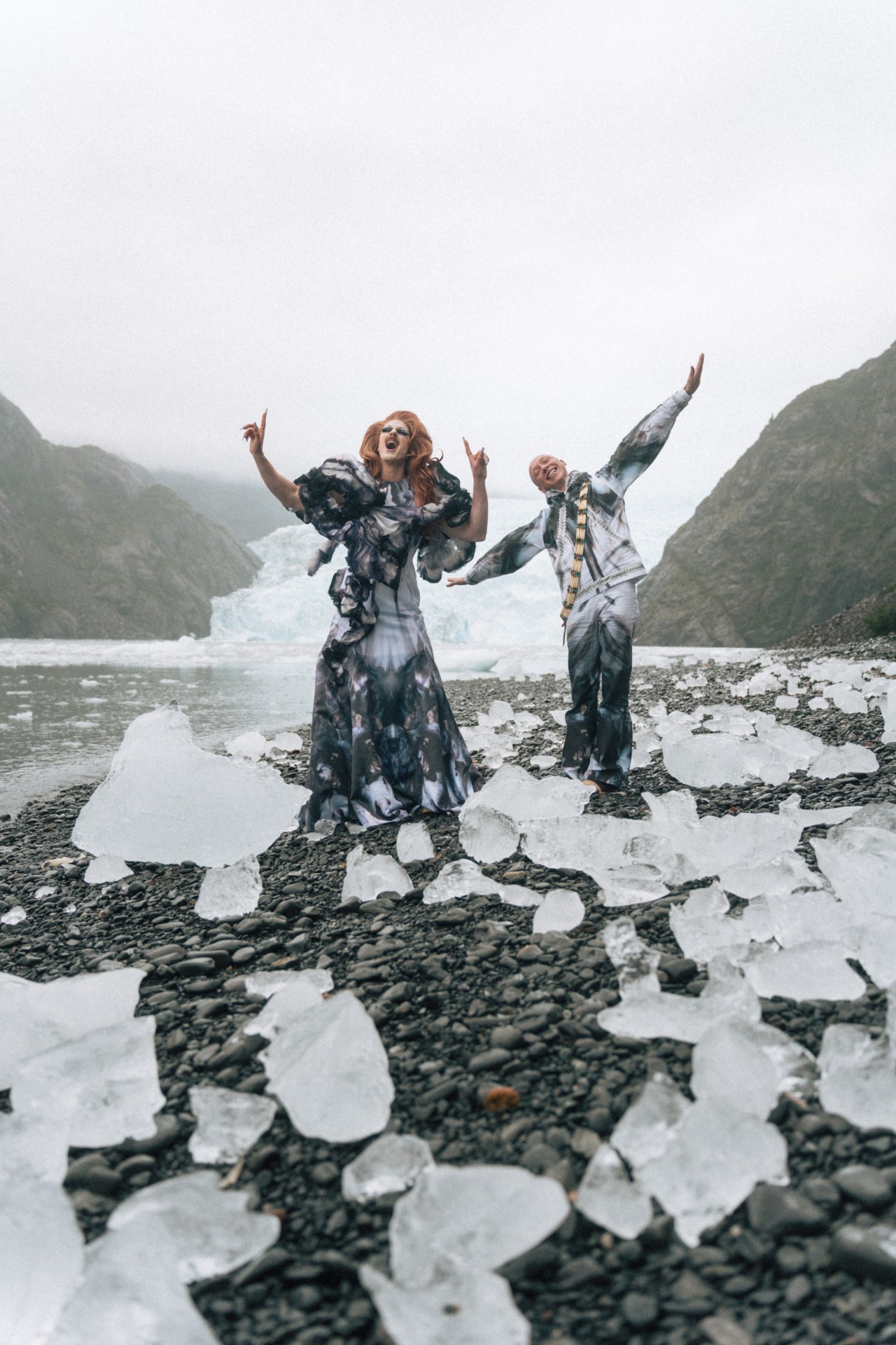 Pattie Gonia and Quinn Christopherson sing while standing on rocky terrain surrounded by blocks of ice.