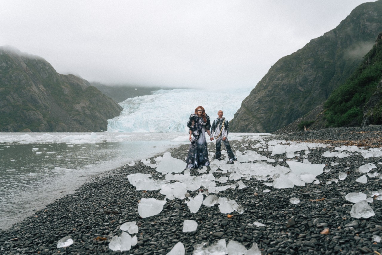 Pattie Gonia and Quinn Christopherson stand among ice and rocks in front of an icy lagoon.