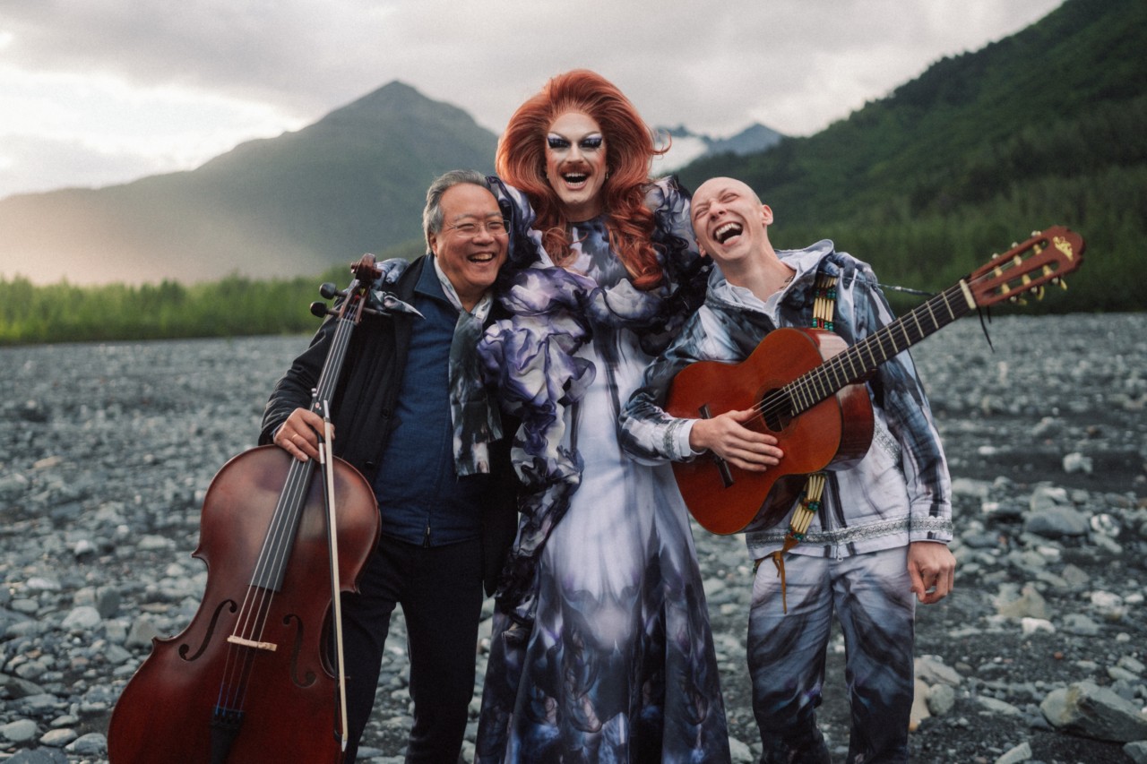 Pattie Gonia, Yo Yo Ma, and Quinn Christopherson pose and smile together in front of a mountain while the sun sets.