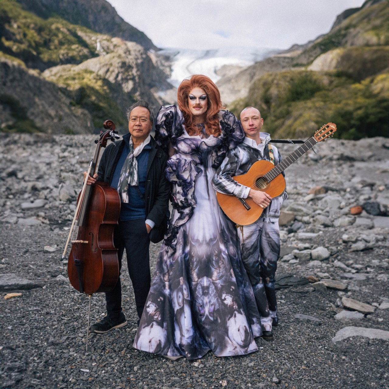 Pattie Gonia, Yo Yo Ma, and Quinn Christopherson pose together with their instruments in front of a rocky, mountainous landscape.
