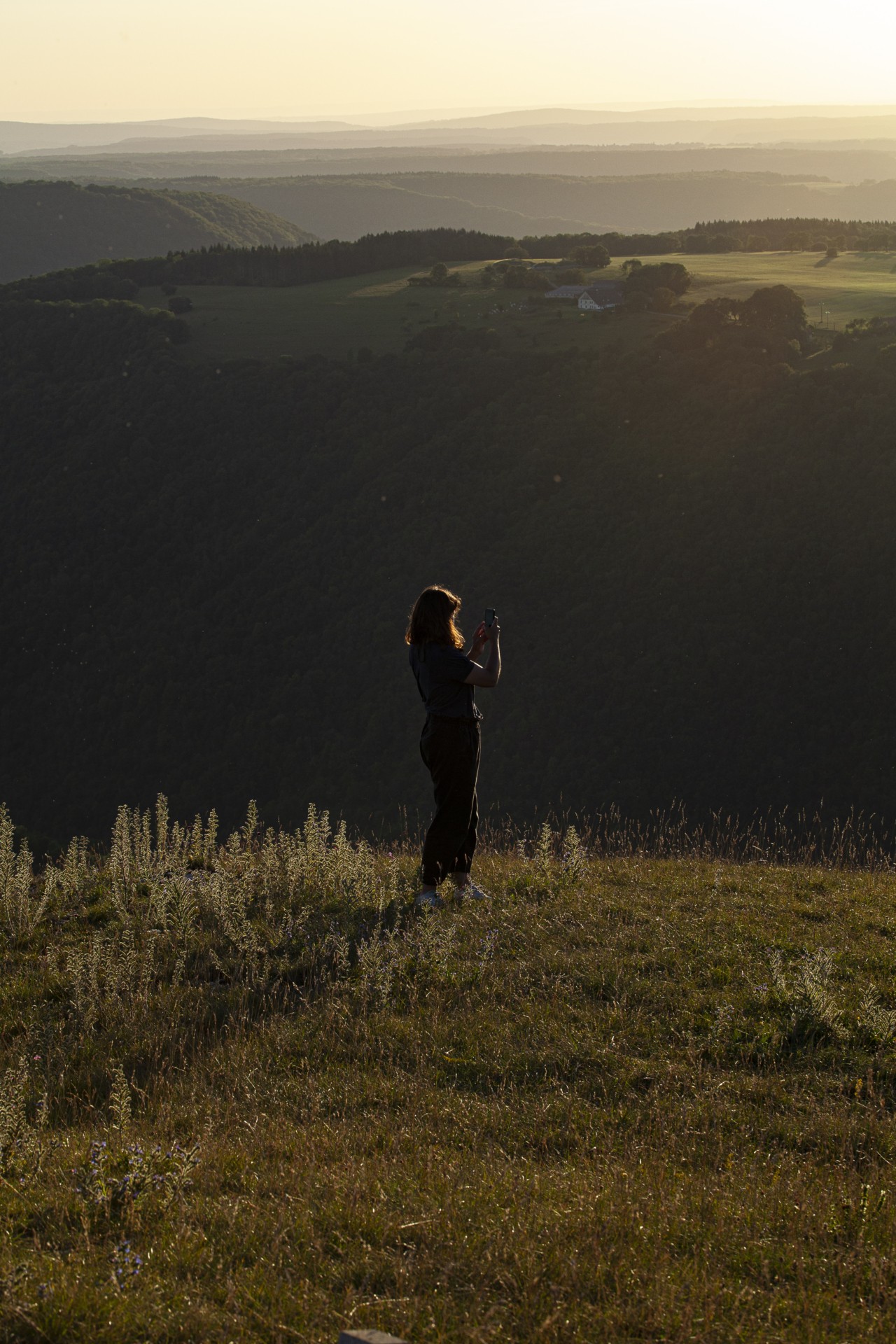 A woman takes a photo while standing near the edge of a grassy, hilly, canyon.