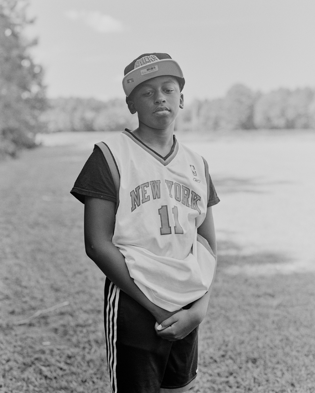 A young boy wearing a jersey and a hat poses in front of a grass field.