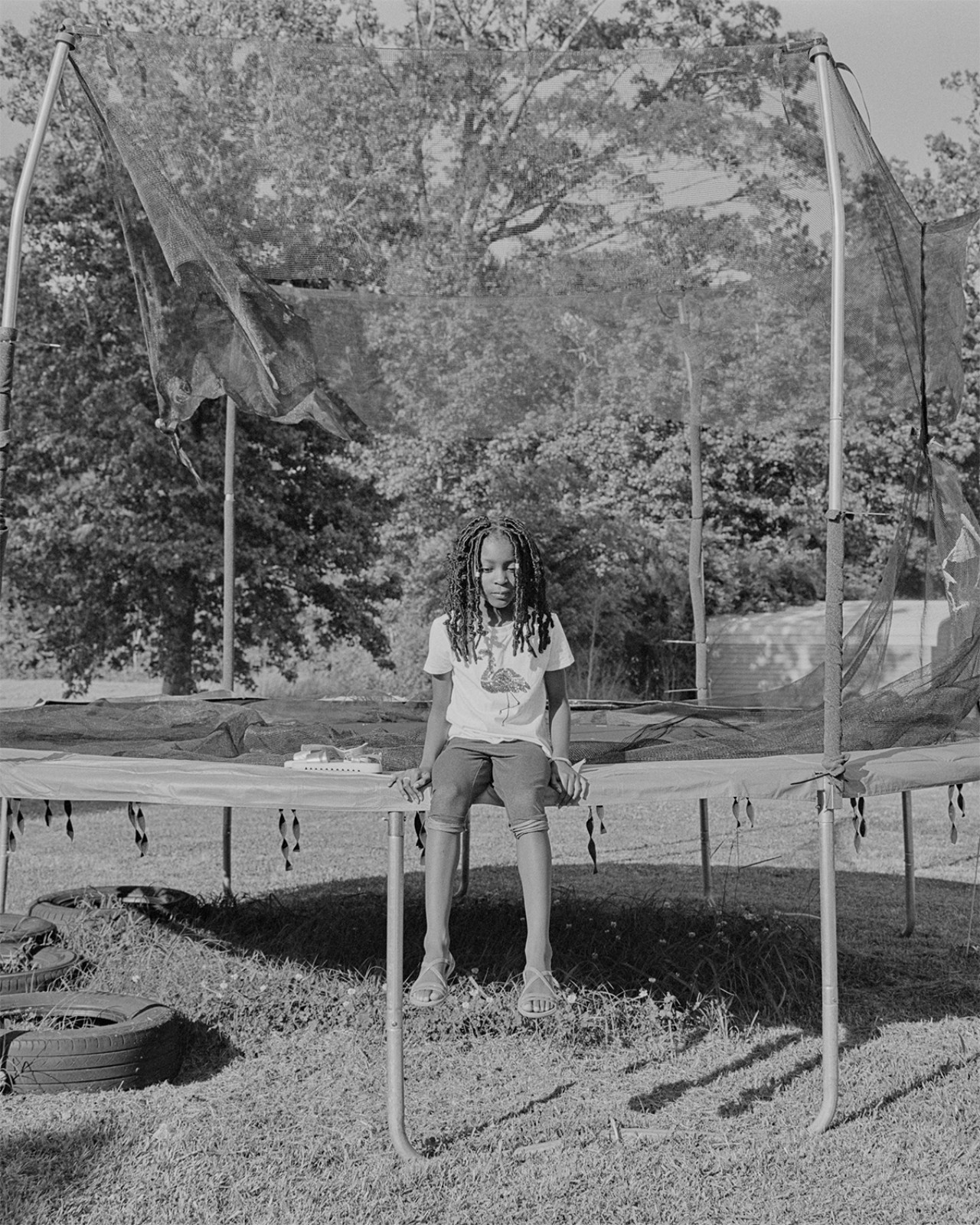 A young girl sits on the edge of a trampoline in a grass field.