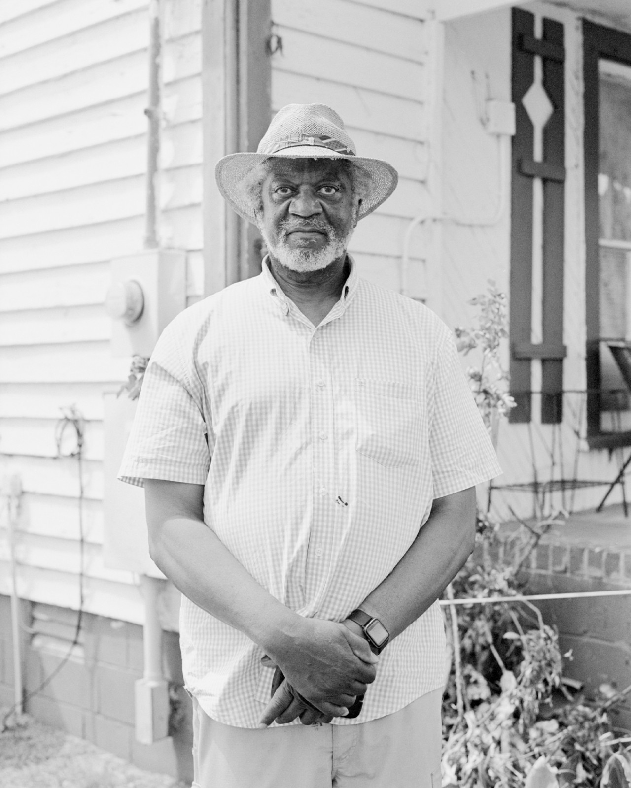 An older man wearing a hat poses in front of a house with this stands clasped in front of him.