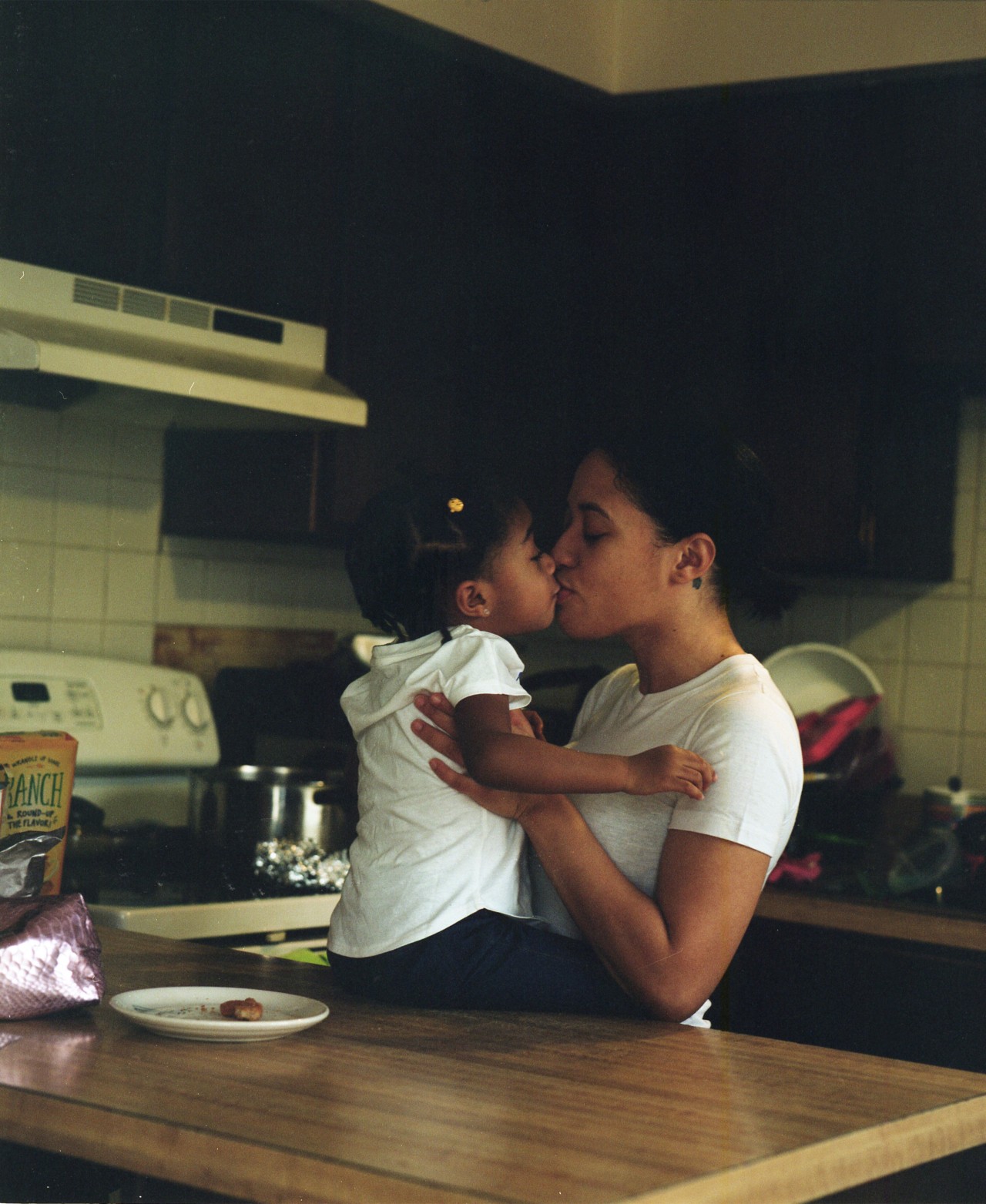 A young mother kisses her child who is propped up on the kitchen table.