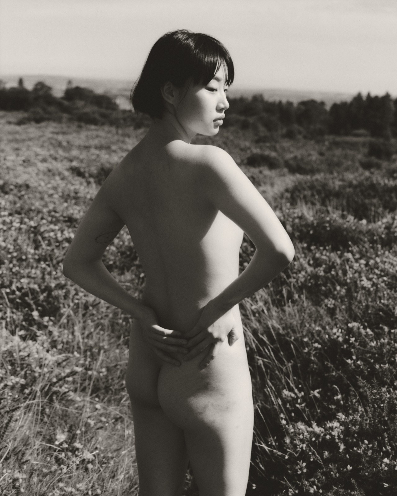 A nude model stands with their hands resting on their back as they face away from the camera.