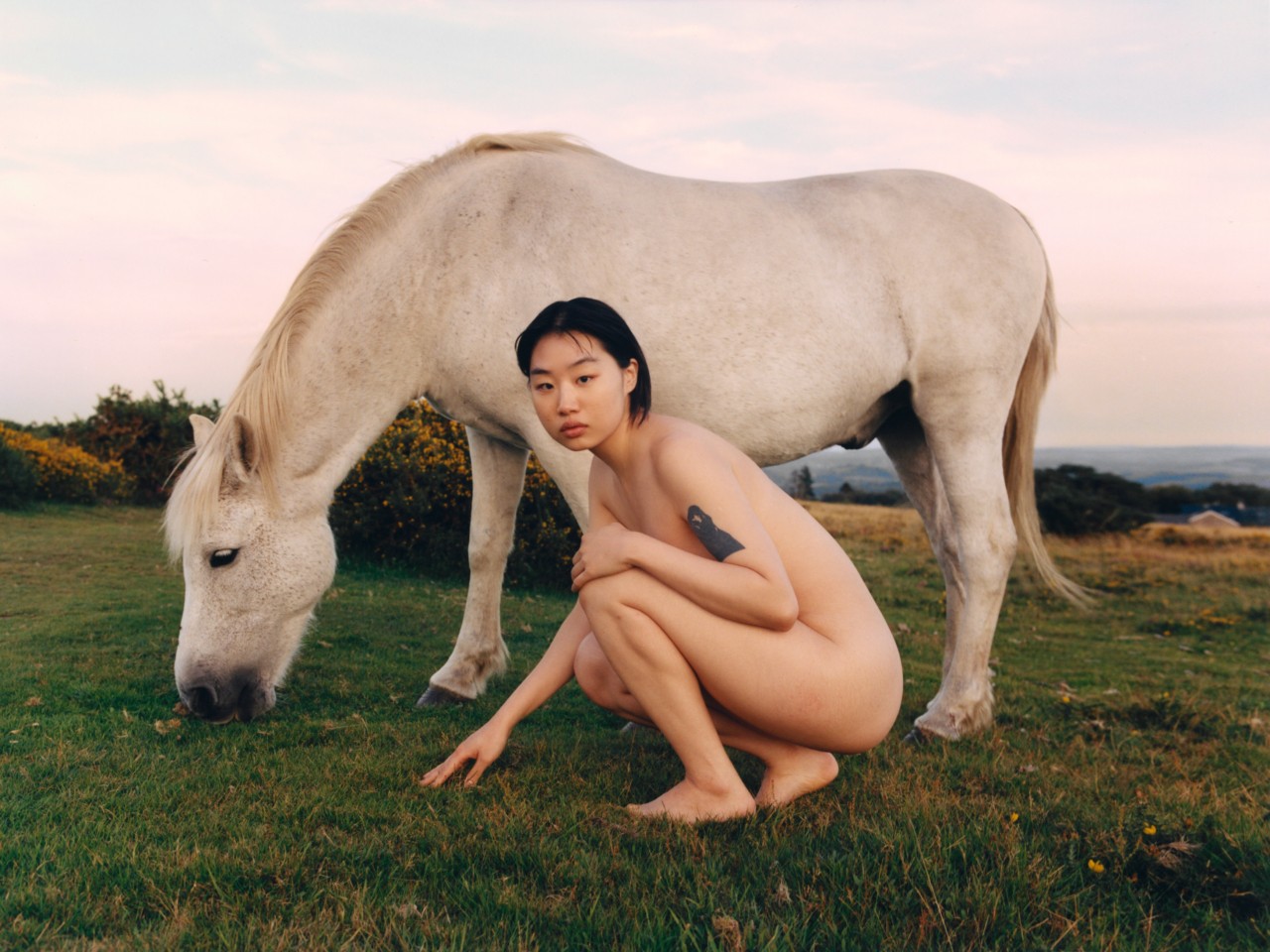 A model crouches on the grass in front of a white horse.
