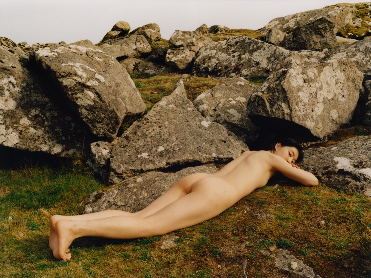 A model lies down along the grass in front of a collection of boulders.