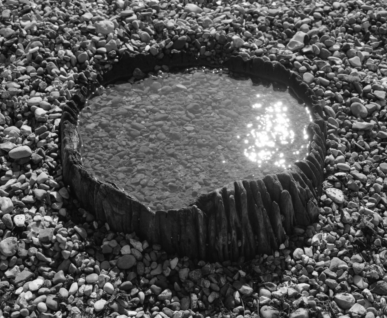 A pool of water encased in a wooden circle sits on a bed of gray rocks.