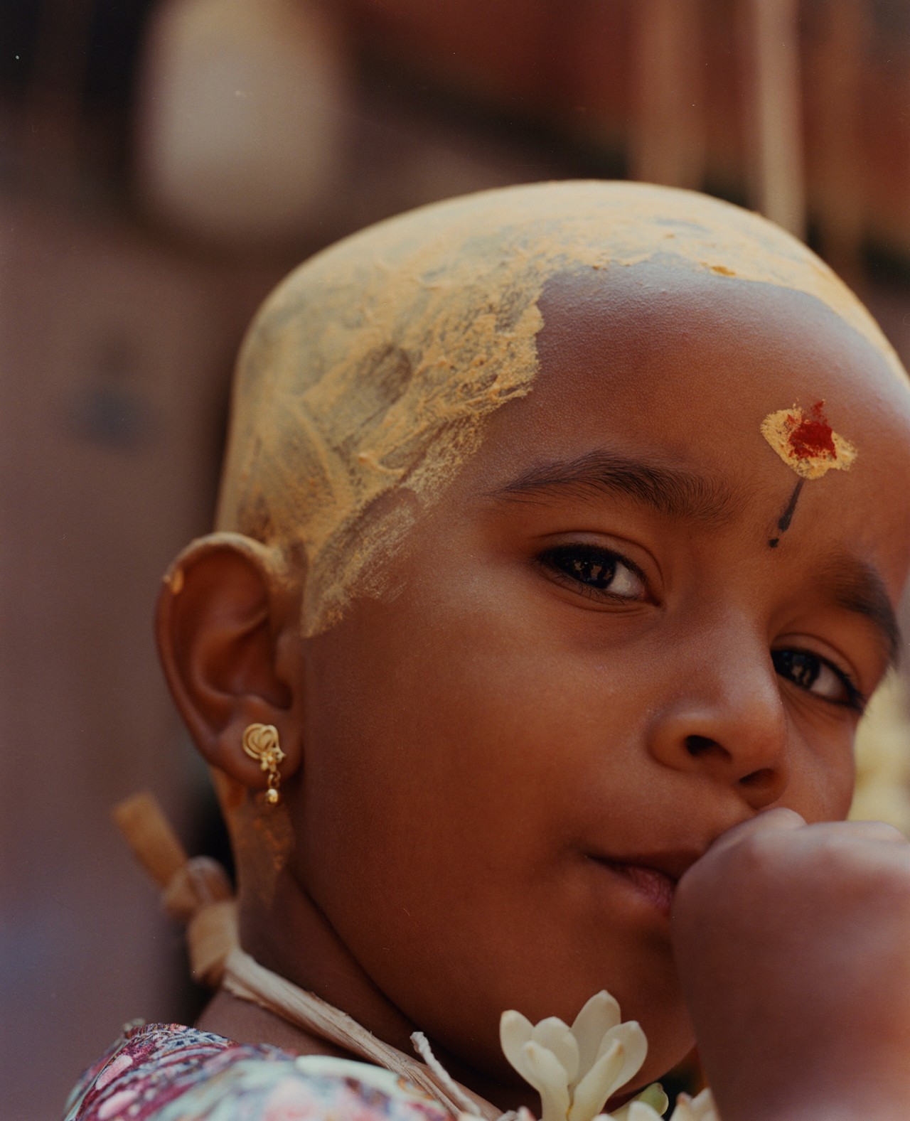 A young boy in Southern India peers at the camera with his thumb in his mouth. The top of his head is painted yellow.