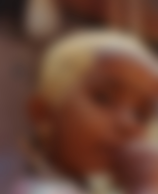 A young boy in Southern India peers at the camera with his thumb in his mouth. The top of his head is painted yellow.
