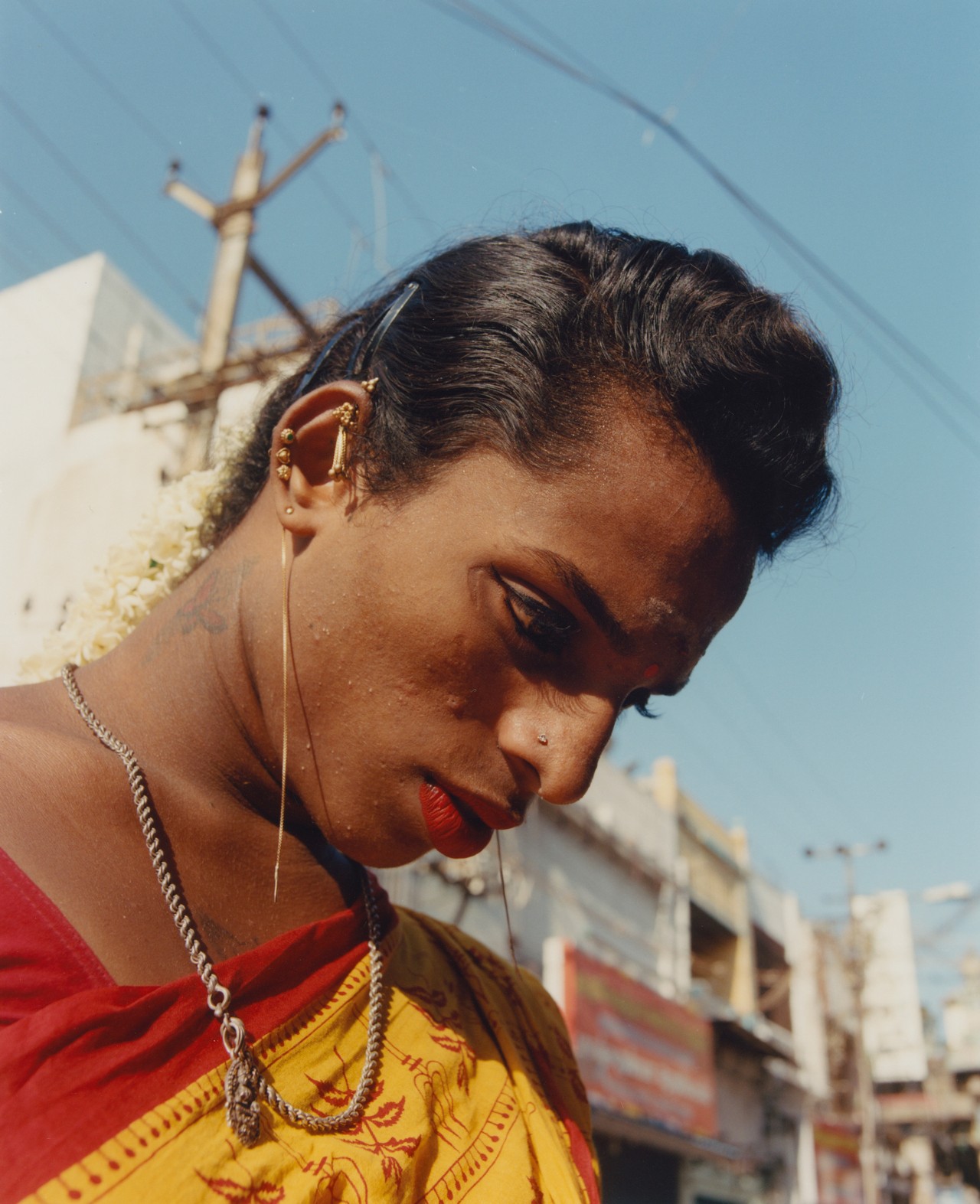 An Indian person with short hair, dressed in traditional red and yellow garb and red lipstick stares down at the ground.