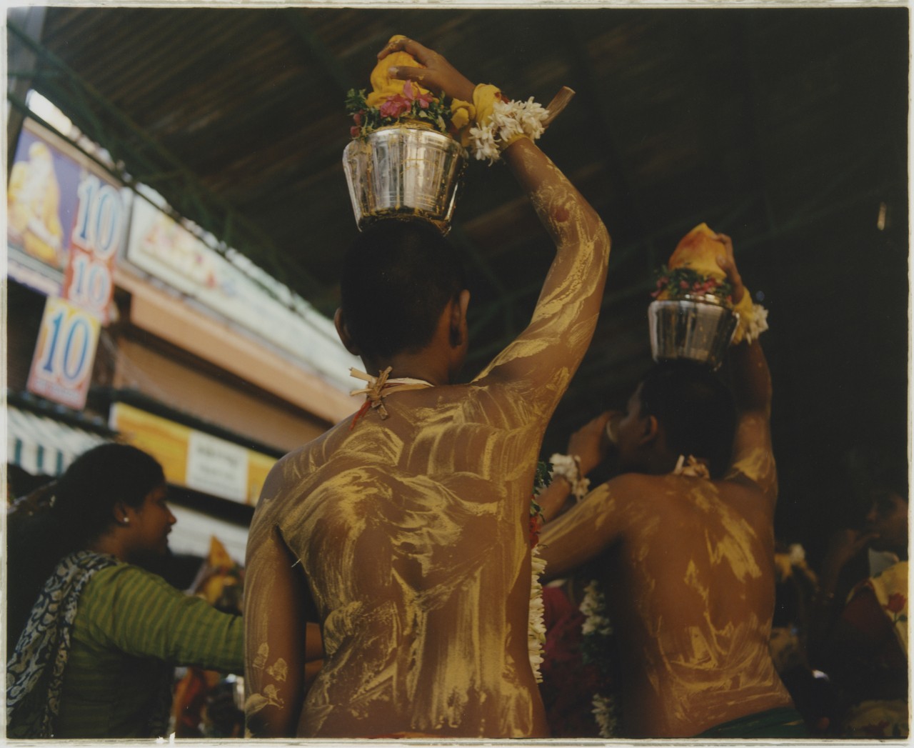 Two young Indian men are covered in dry yellow paint as they walk holding a filled silver container over their head with their right hand.