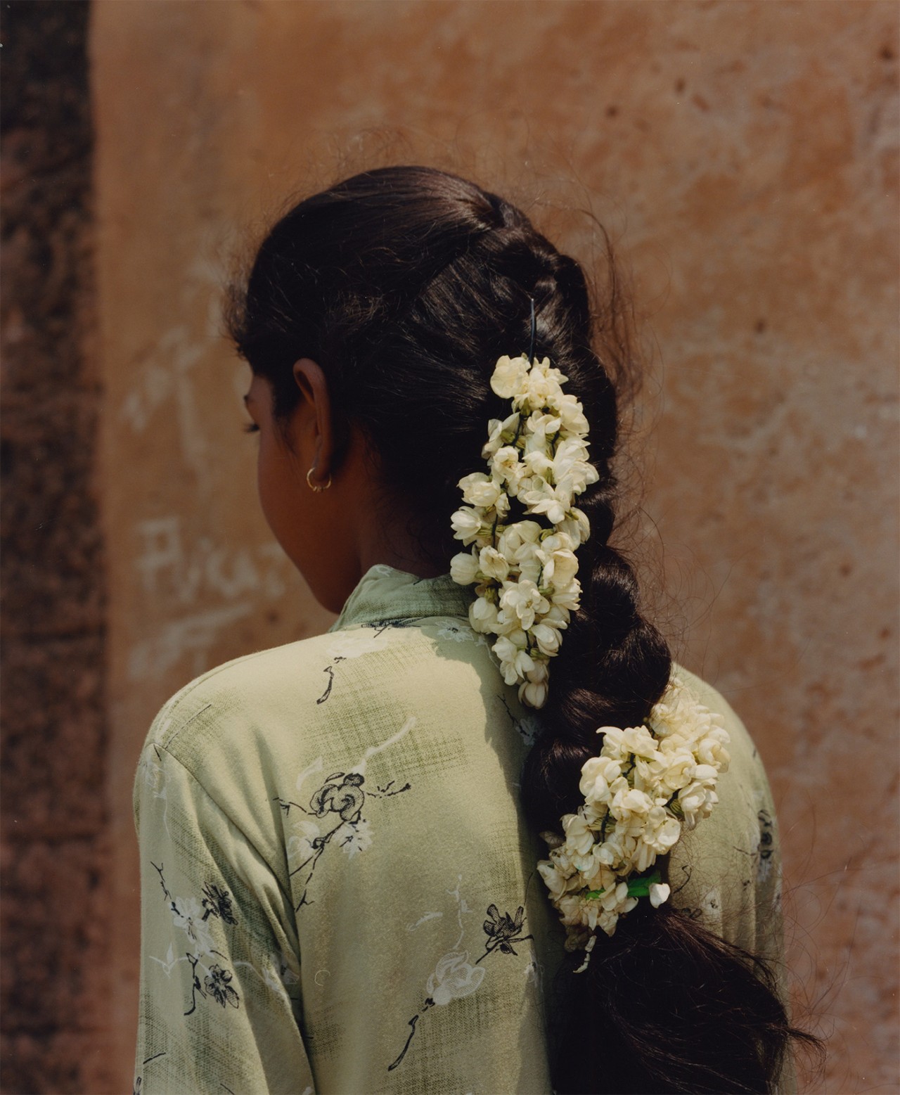 A young Indian girl faces away from the camera. Her hair is gathered into a large braid ring down her back with white flowers weaved into it.