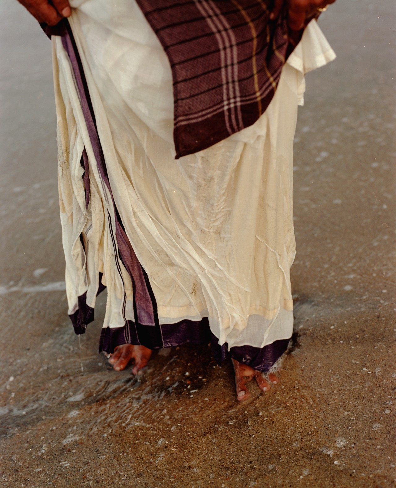 A person wearing traditional garb stands on the shore of a beach. Their feet and the bottom of their clothes are wet from the water.