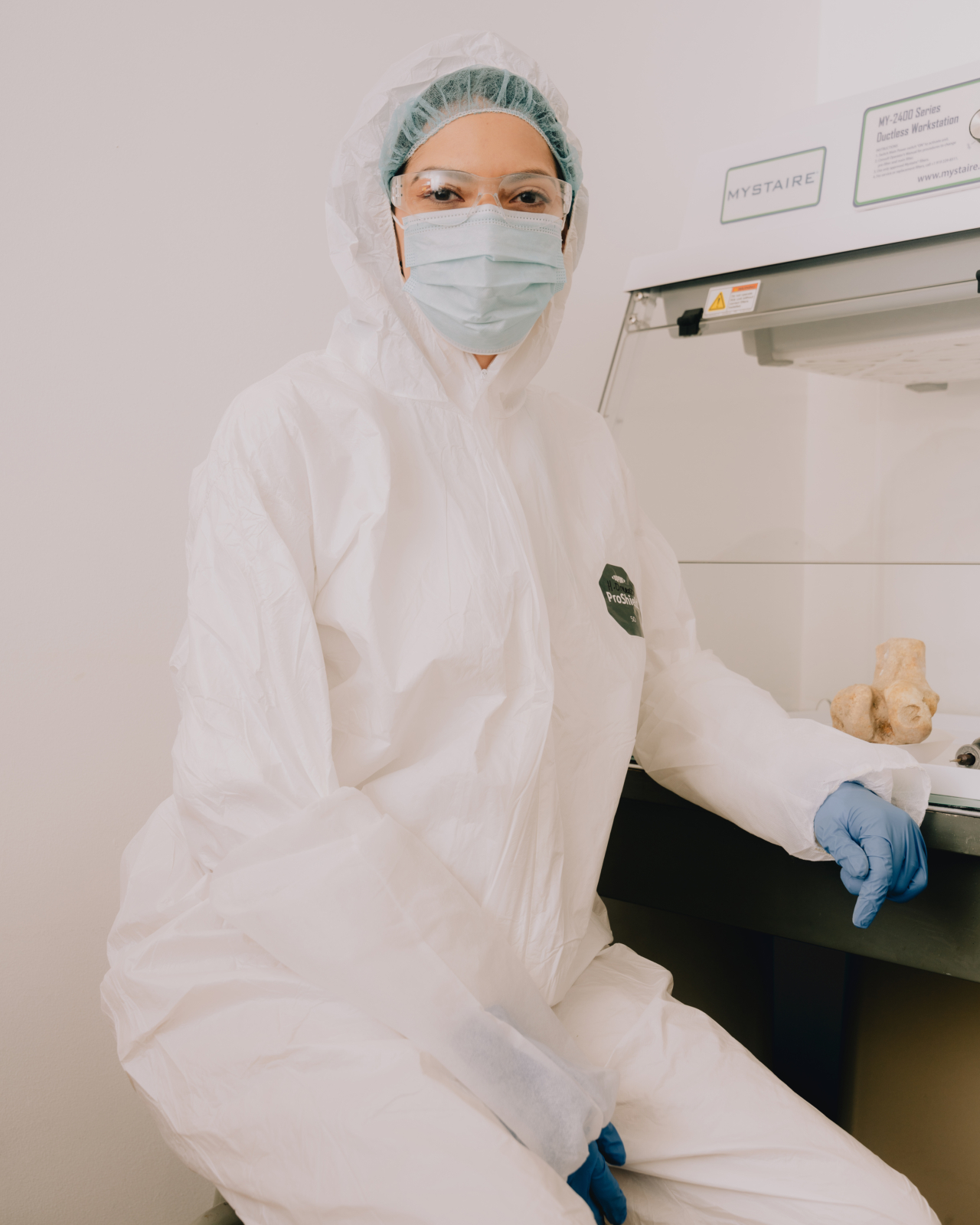A scientist wearing a mask, protective eyewear, and a suit sits on a stool in a forensics lab.