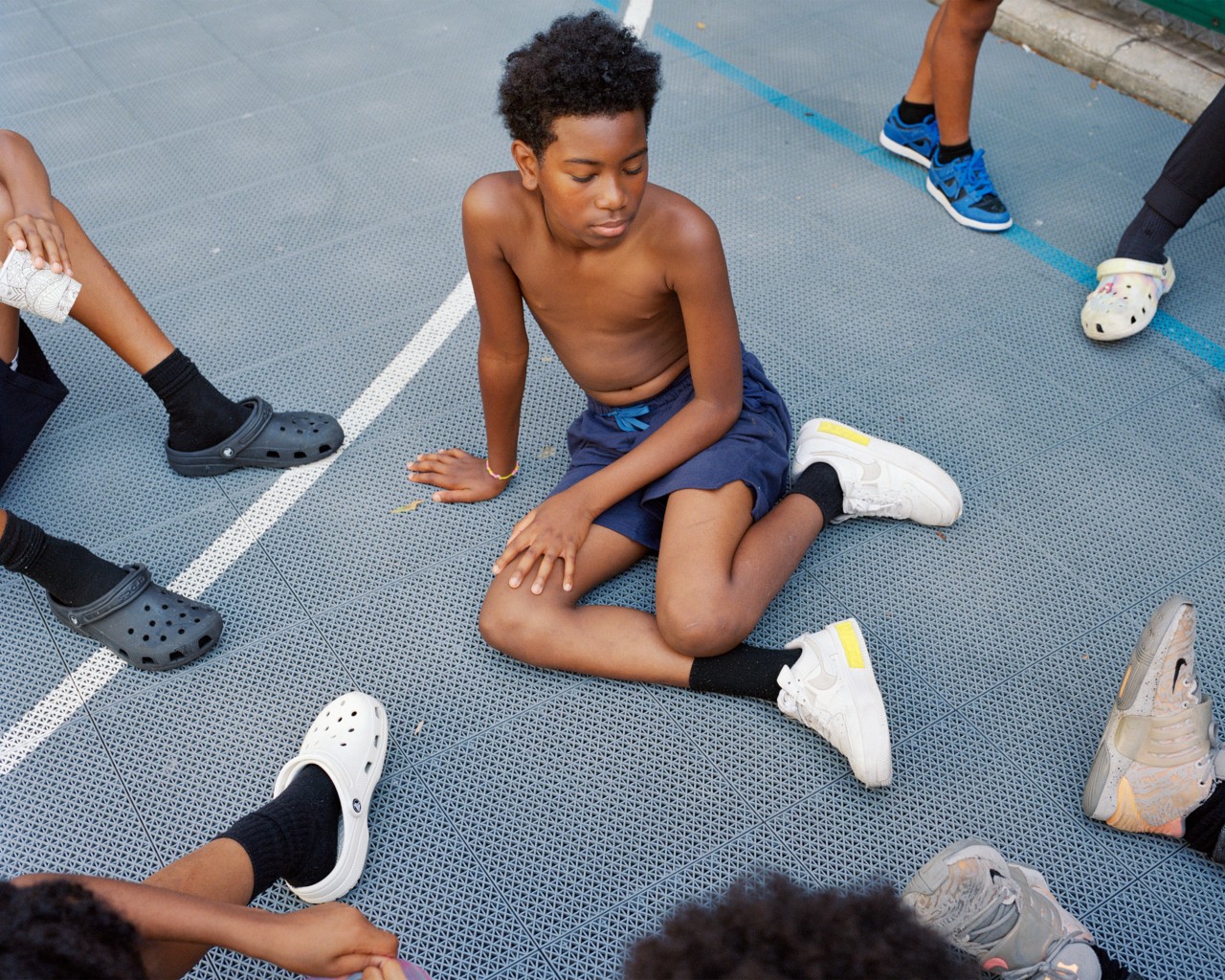 A boy sits outside among his peers on the ground of a basketball court.