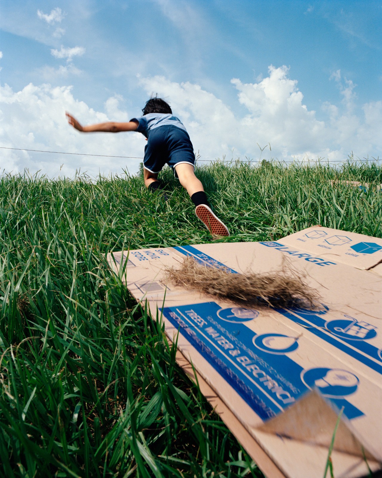 A child runs up a grass hill. A flattened cardboard box lays flat on the grass.
