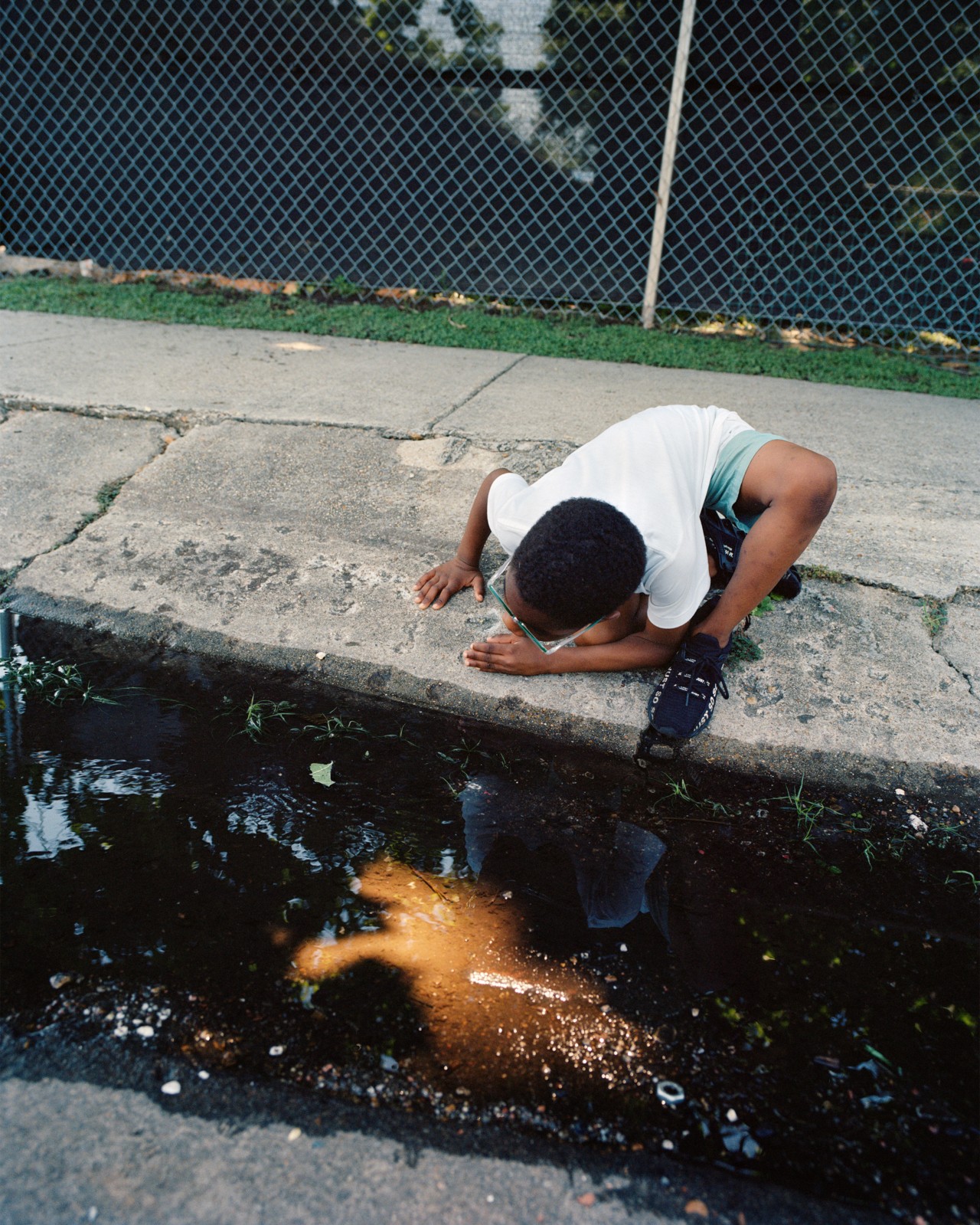 A young boy crouches on the sidewalk of New Orleans examining the reflective light in a street puddle.