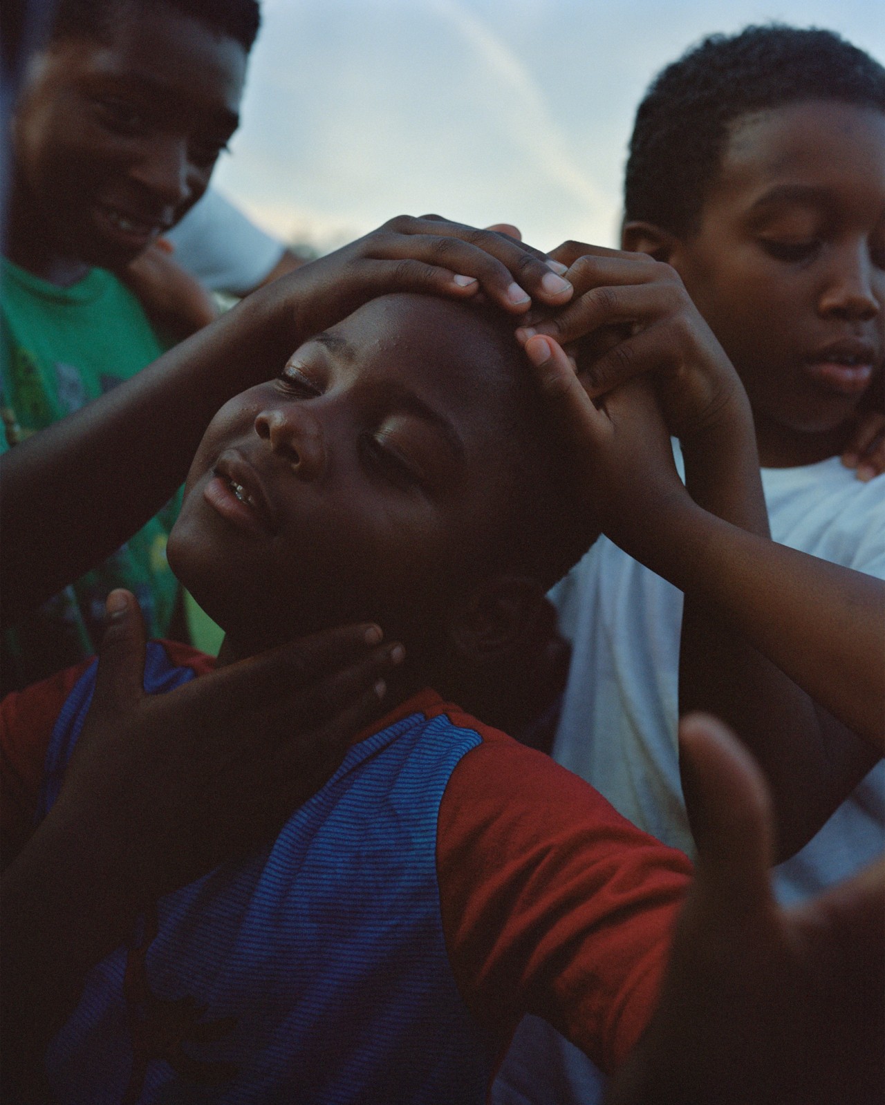 Several boys surround a younger boy, placing their hands on top of his head.