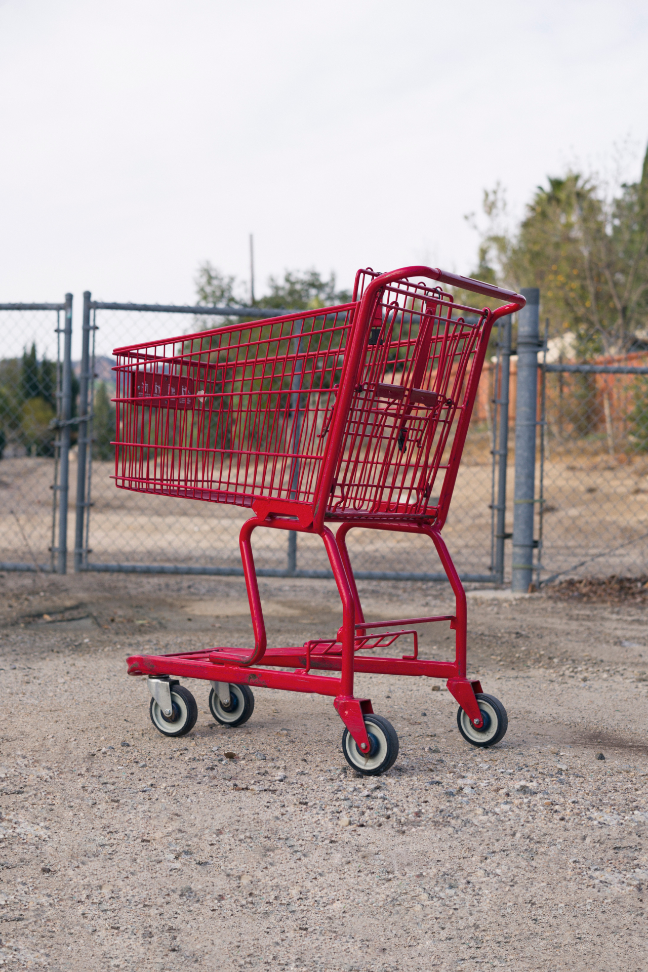 A lone, red shopping cart is placed in front of a metal gate on a dirt path.