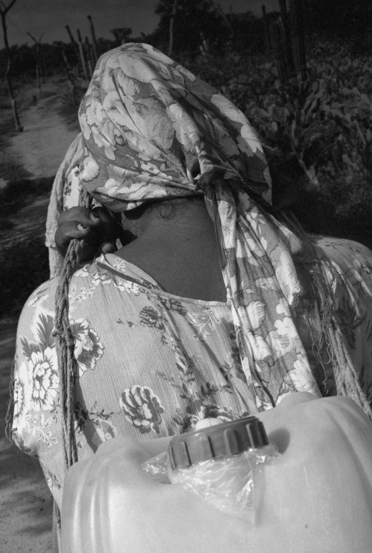 An Indigenous person wearing a scarf and dress carries an empty water jug as they face away from the camera.