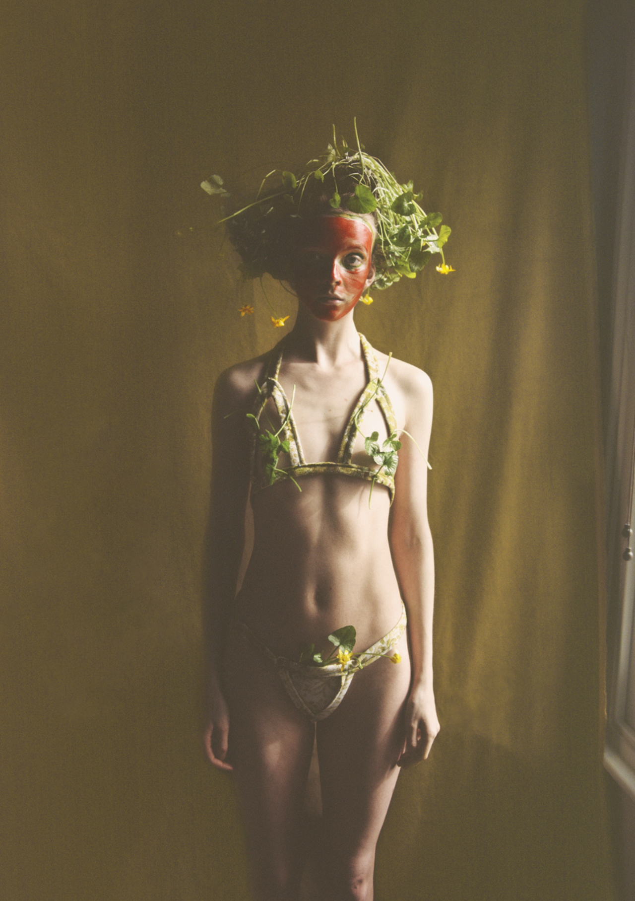 A model wears red paint on their face and a bikini and headpiece made from plants.