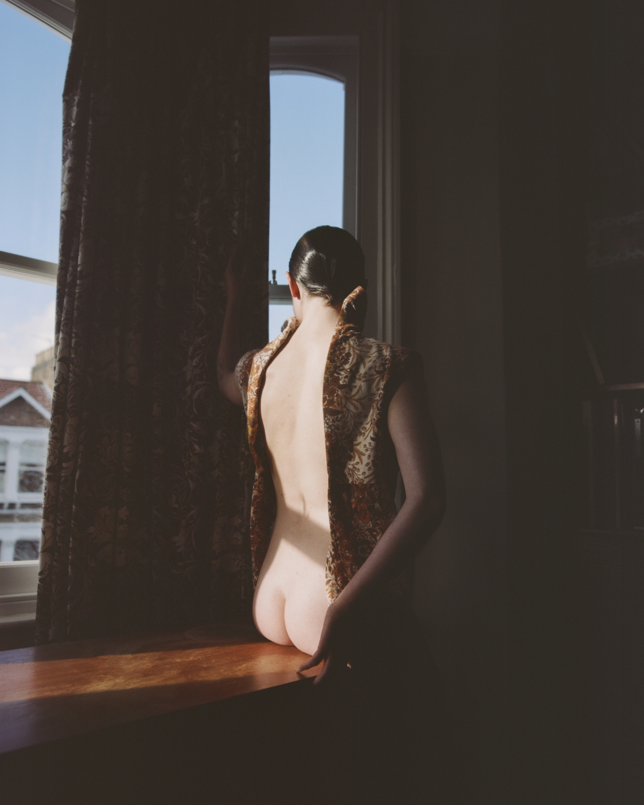 A model sits on a desk while facing out a window with sunlight peering through.