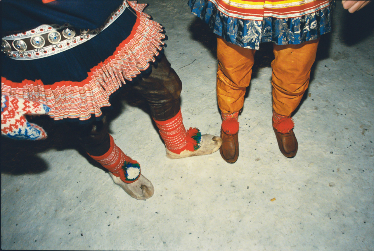 Two Sami people stand on the hard snow. One person uses their foot to touch the other person's foot.