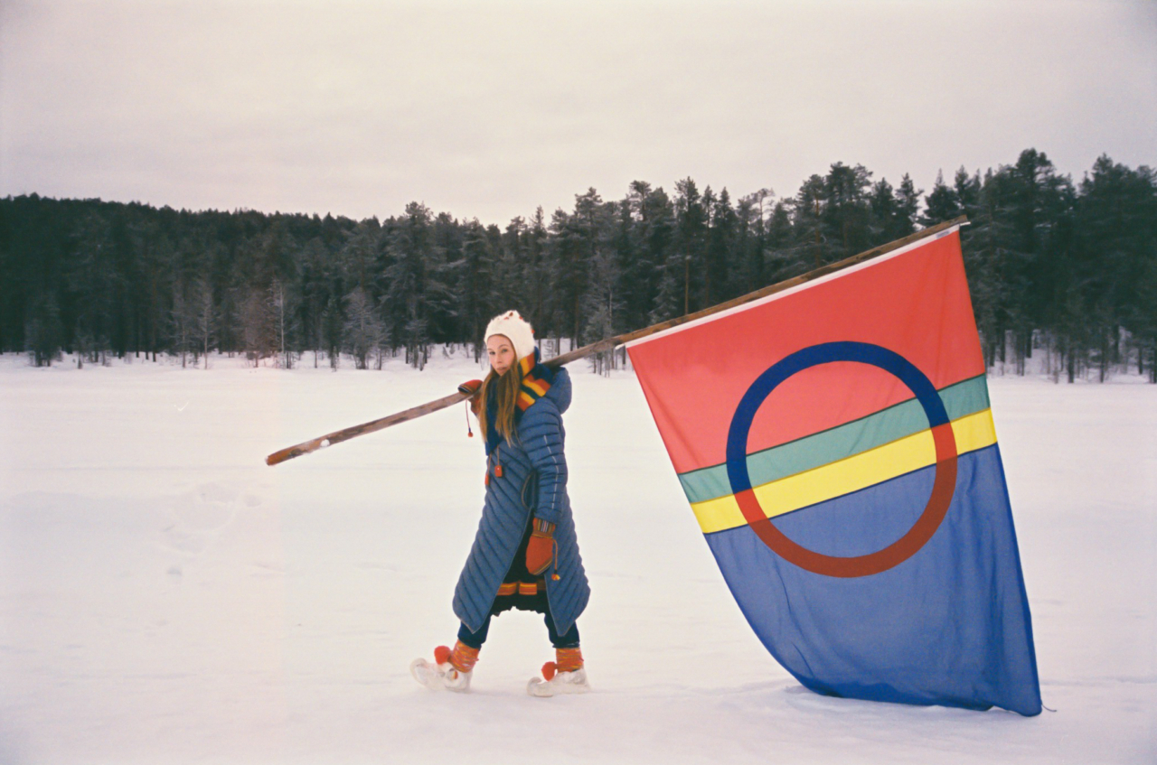 Sofia Jannok carries a large flag through the snowy clearing.