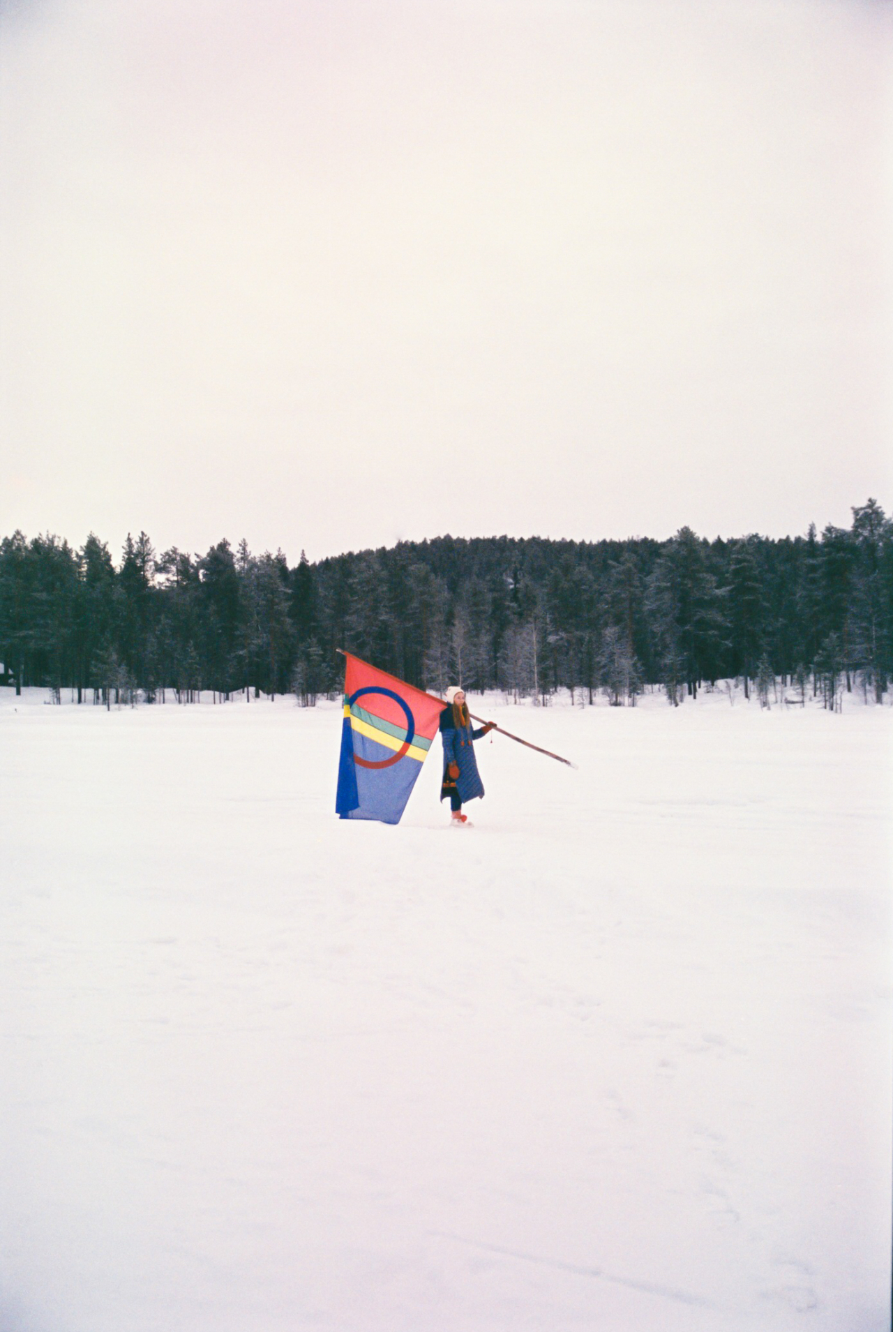 A Sami person stands in the middle of a snowy forest holding a colorful flag.