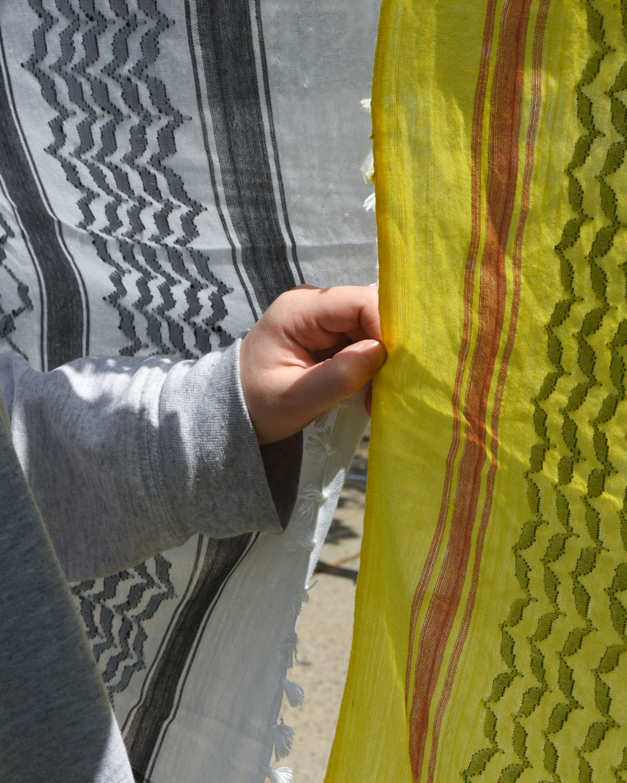 A hand holds a hanging yellow, black, and red keffiyah. A black and white one hangs right next to it.