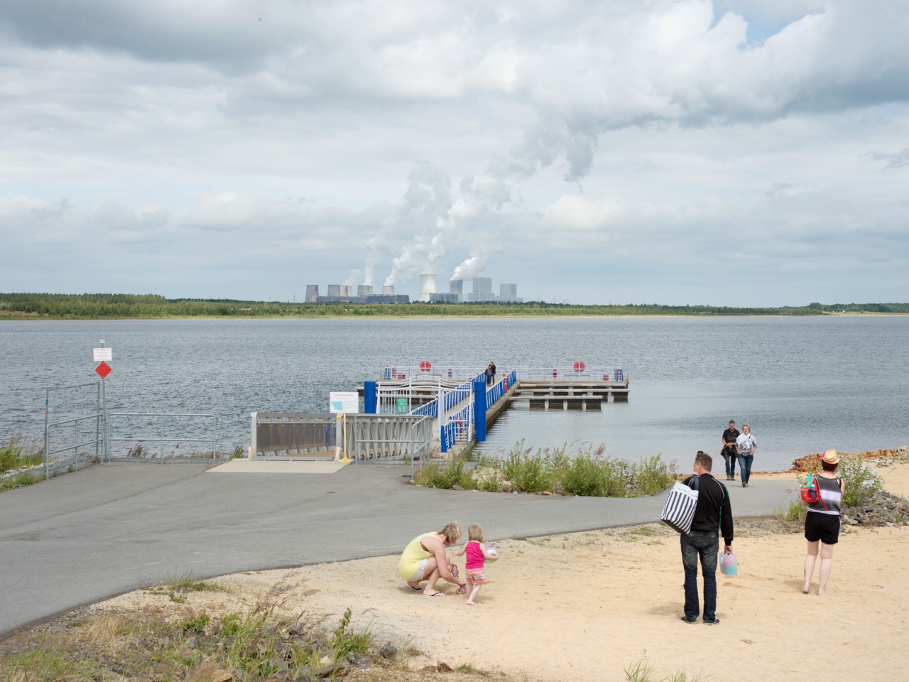 People gather near a dock on a beach. On the other side of the body of water are factories and coal mines releasing fossil fuels into the air.