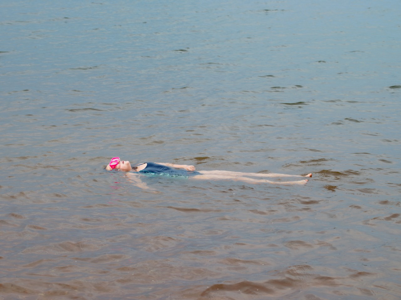 A person wearing a blue one piece swimsuit and pink swim cap floats on their back in the middle of a body of water.