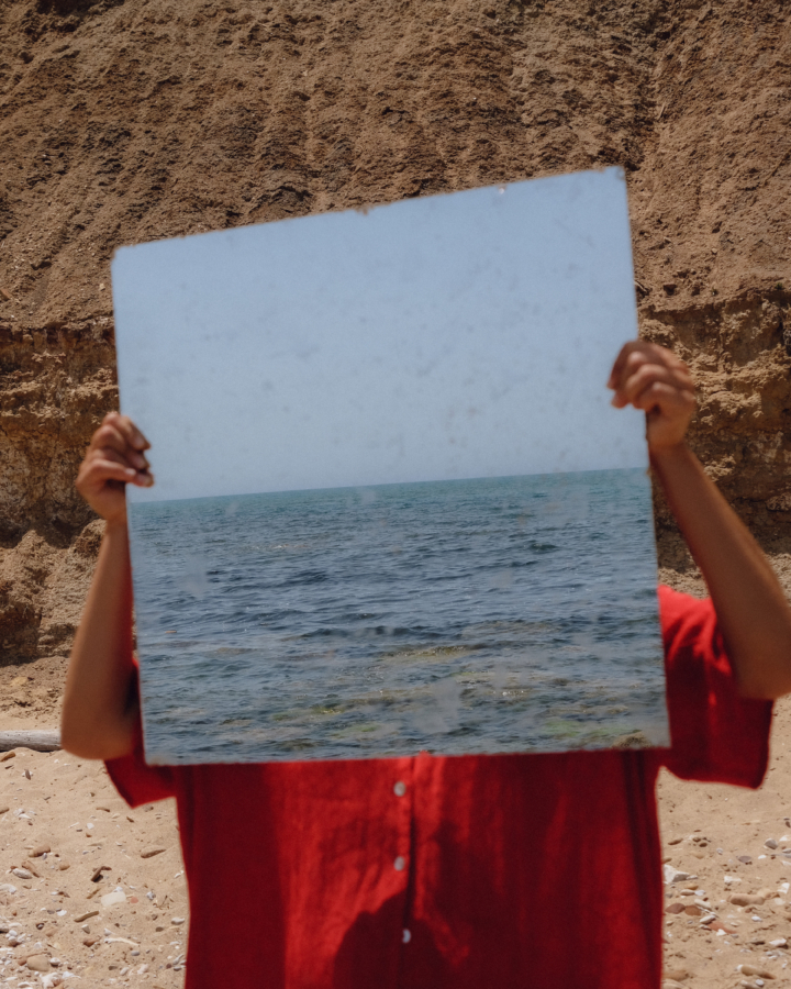 A young person stands in front of a dirt mound wearing a red button up shirt and holds a portrait of an ocean in front of their face.