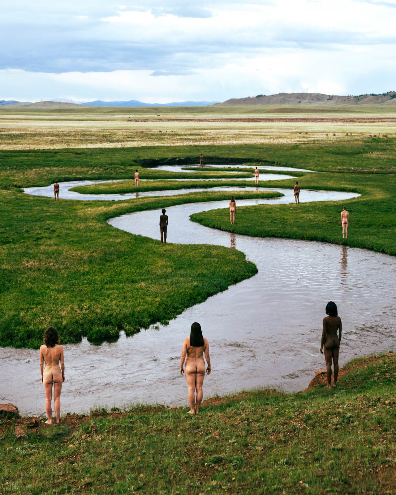 Naked people stand on the grass along a windy river.