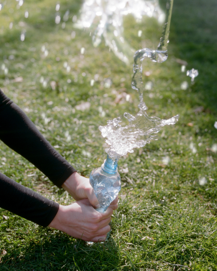 Two hands squeeze a water bottle so that water splashes out of it and on to the grass below.