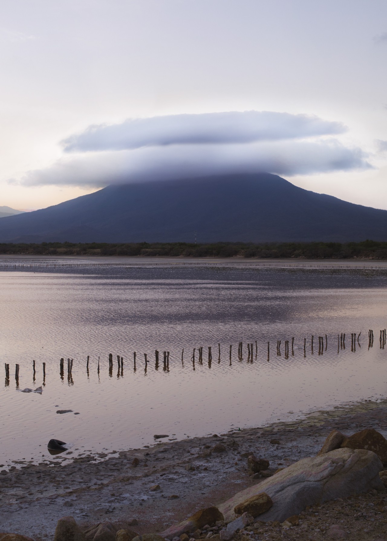 A cirrus cloud hovers above a distant mountain that sits behind a body of water at dusk.
