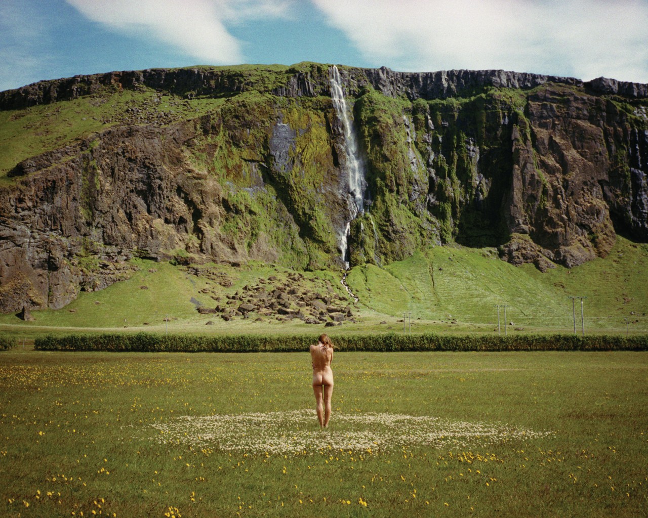 A nude woman stands in the middle of a meadow, facing a grassy cliff with a waterfall.