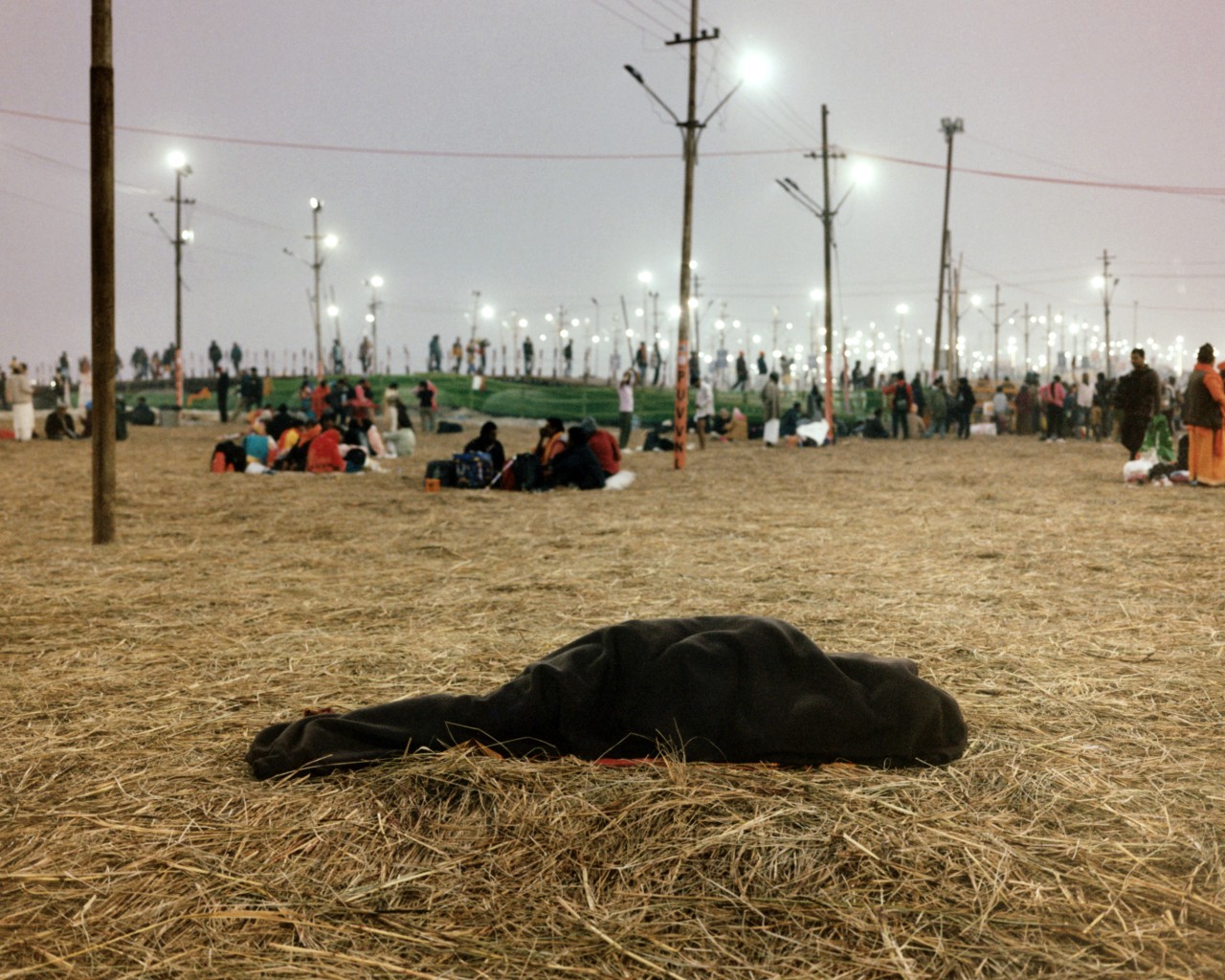 A man rests under a blanket, lying on dry grass laid out for insulation from the cold ground.