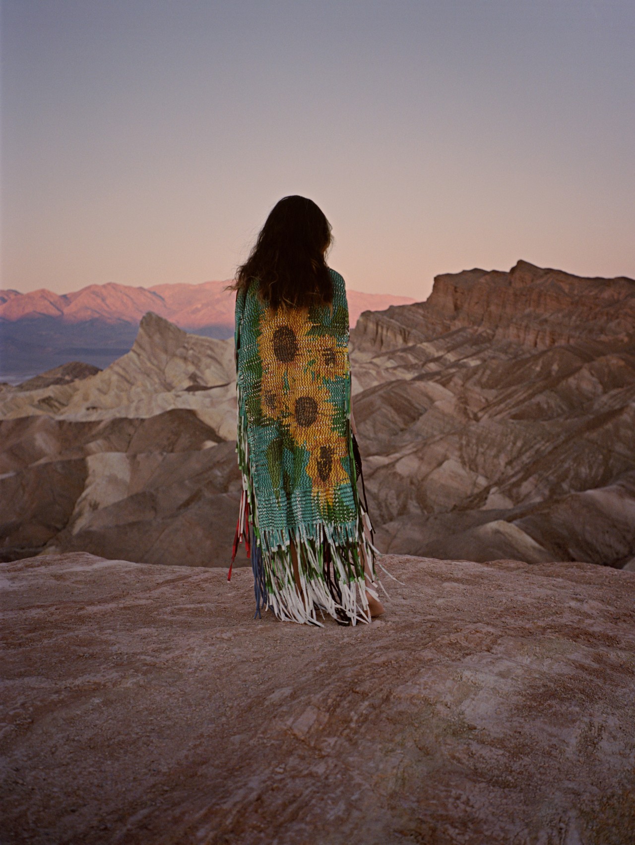 A person with long brown hair wears a tunic with sunflowers on the back as they stand among the mountains.