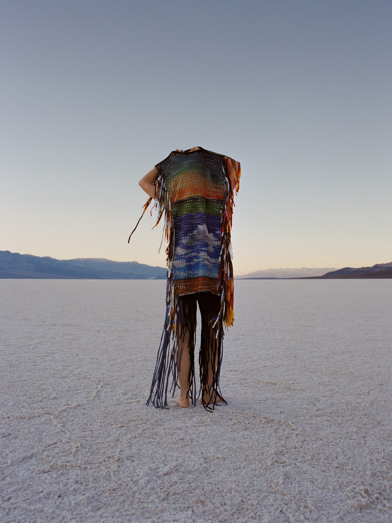 Someone stands in a vast landscape and covers their face and body with a multi-colored tunic.