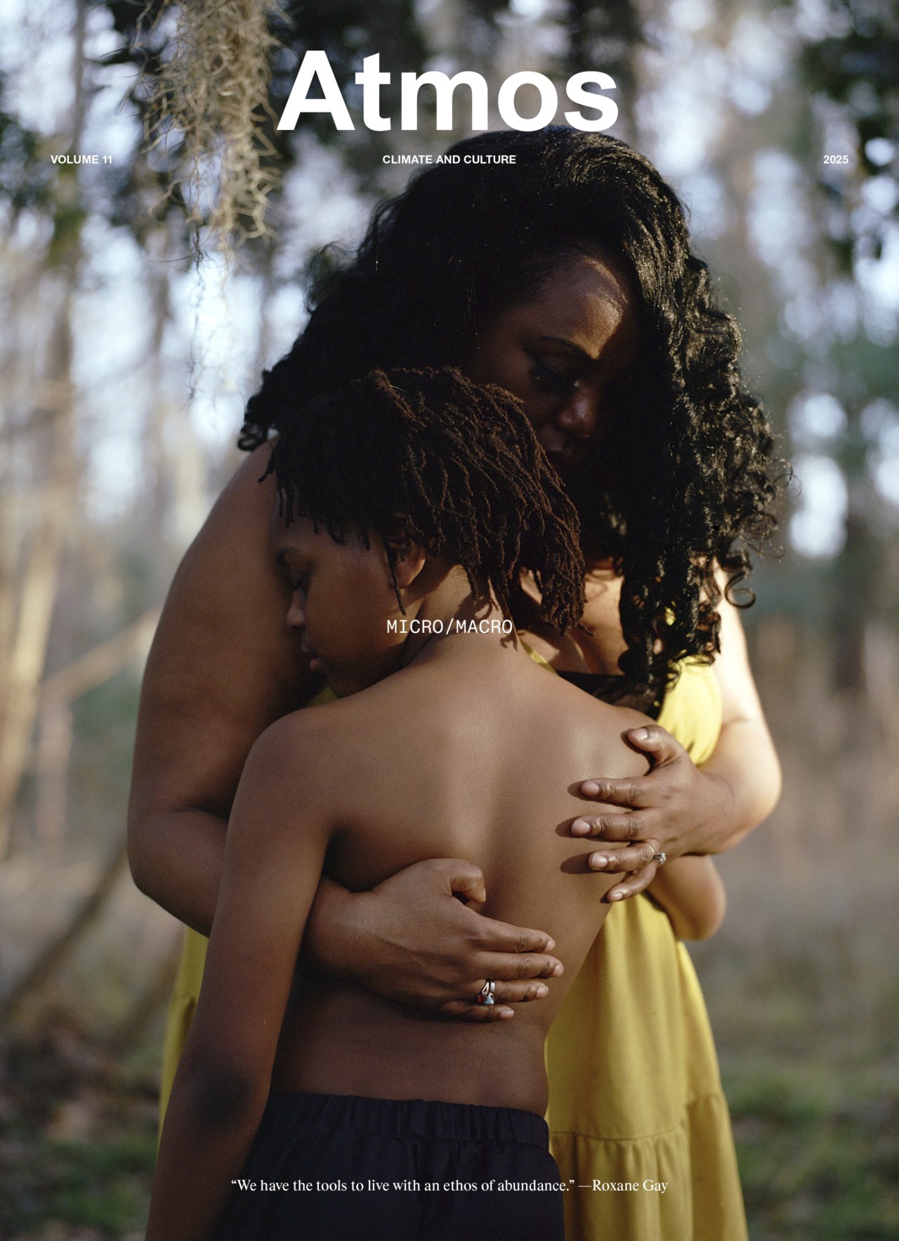 A woman in a yellow dress gently embraces a child outdoors, both with eyes closed, conveying comfort and warmth. The image is featured on the cover of Atmos magazine, with trees blurred in the background.