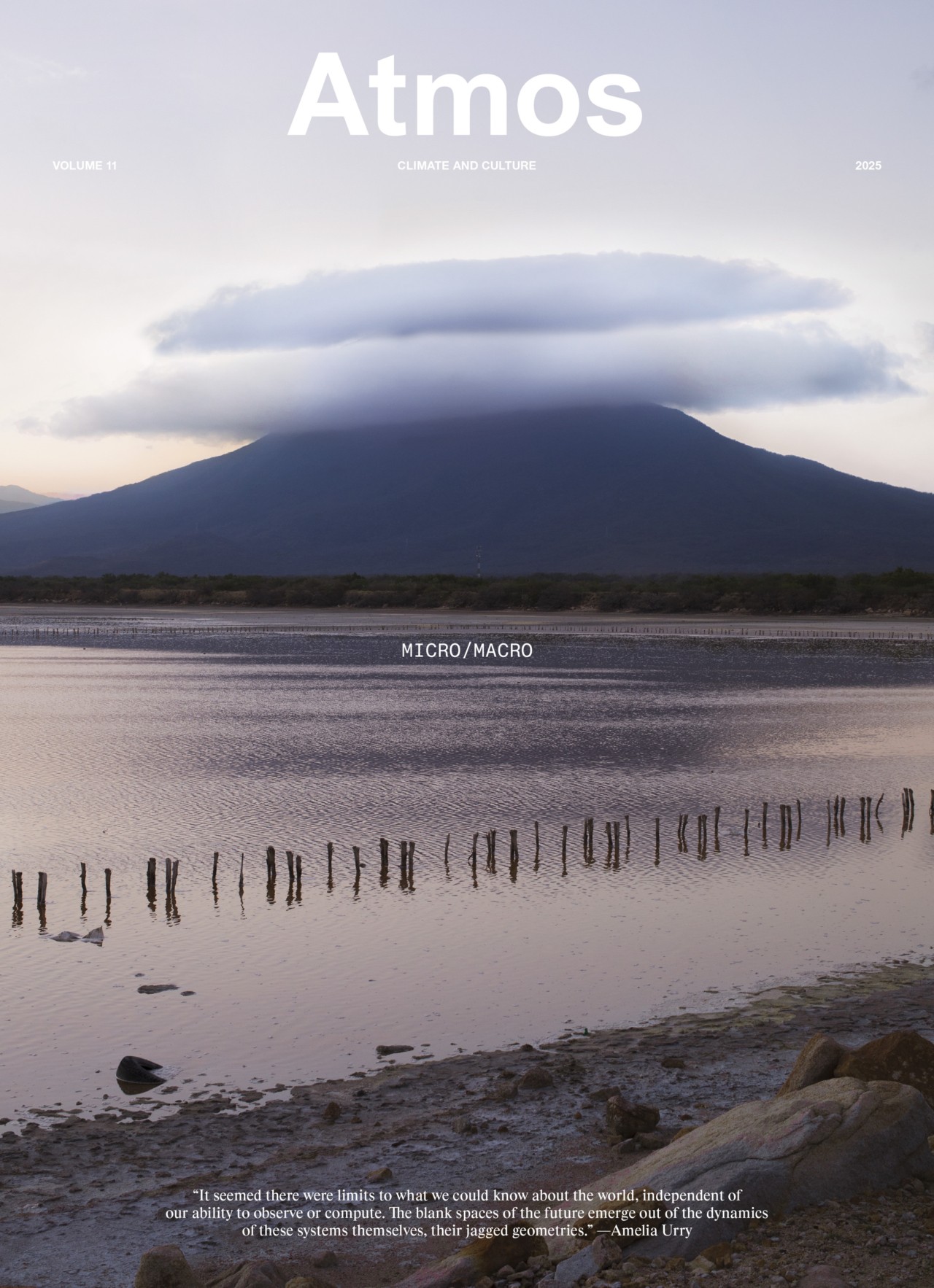 A tranquil lake with old wooden posts, rocky shore, and a misty, lenticular cloud-capped mountain in the background at dusk; the scene is featured on the cover of Atmos magazine, Volume 11, titled Micro/Macro.