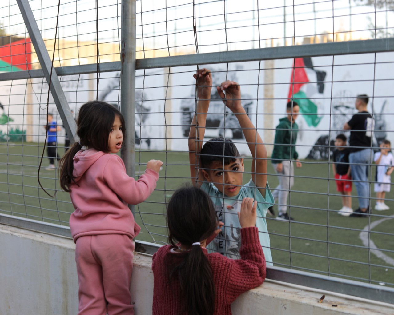 Palestinian children stand next to the gate of a soccer field.