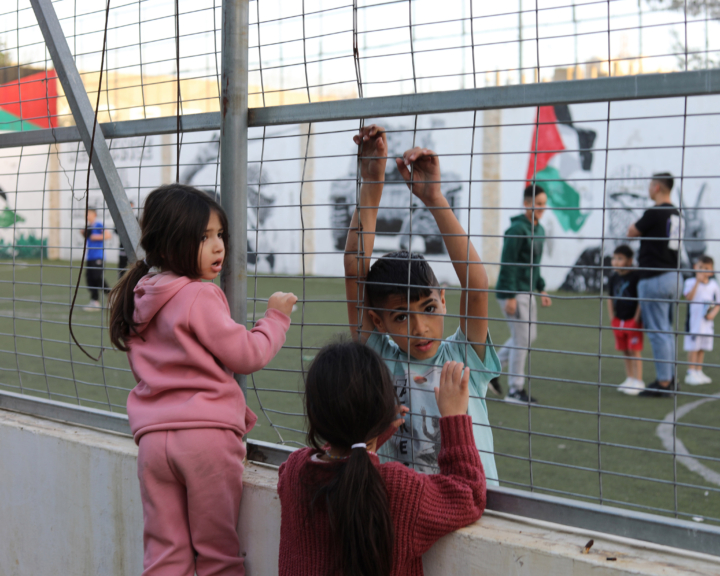 Palestinian children stand next to the gate of a soccer field.