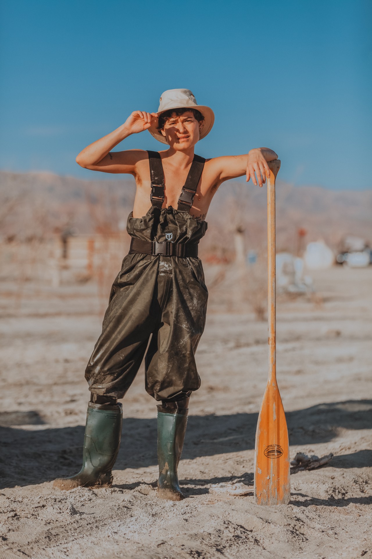 A person wearing overalls, a hat, and boots leans against a paddle while standing on the shore of the Salton sea.